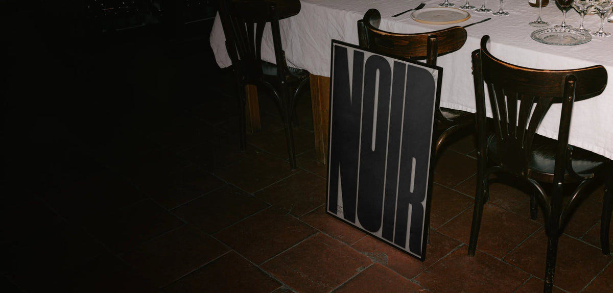 Noir poster by Meinhard Taxer in a black frame, leaning against a chair in a dark restaurant